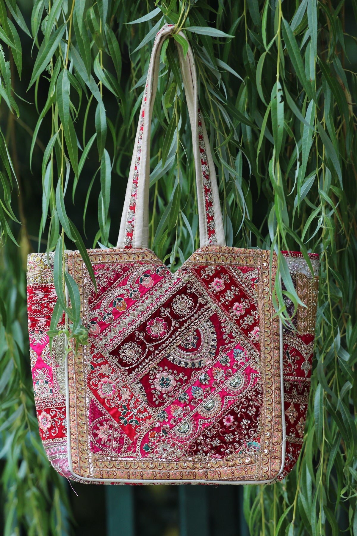 A large embroidered tote bag with red and pink patterned fabric and gold metallic embellishments, hanging in front of green willow leaves.