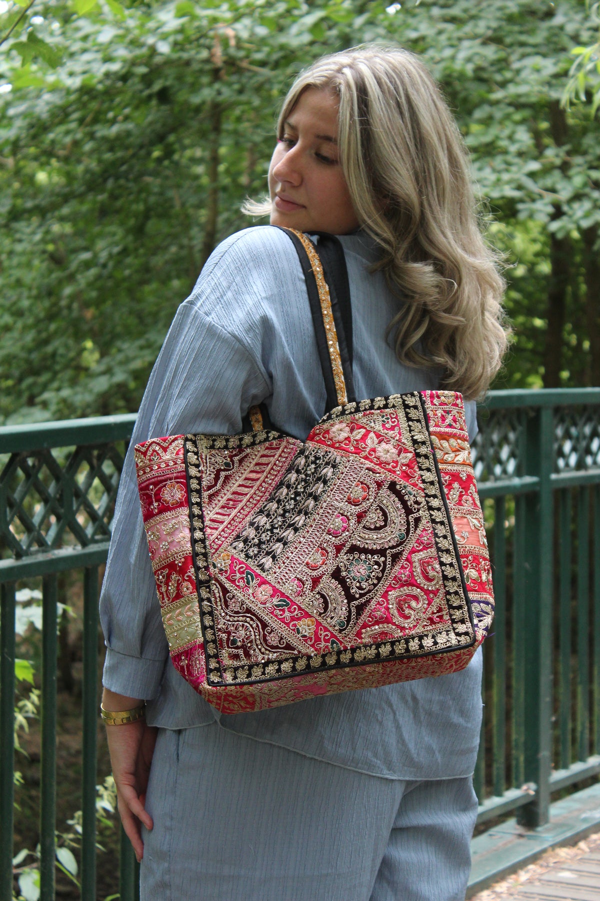 A woman wearing a blue denim outfit carries a large, vibrant bohemian-style tote bag with intricate red, pink, black, and gold patterns, set against a green outdoor background.