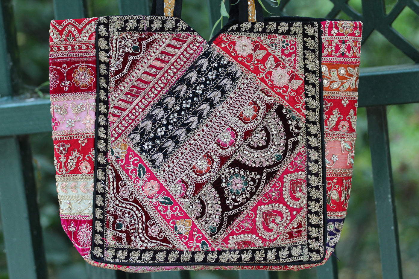 A handmade patchwork tote bag made from multiple red, pink, black, and beige patterned fabric panels with ornate embroidery, hanging against a dark green metal fence.