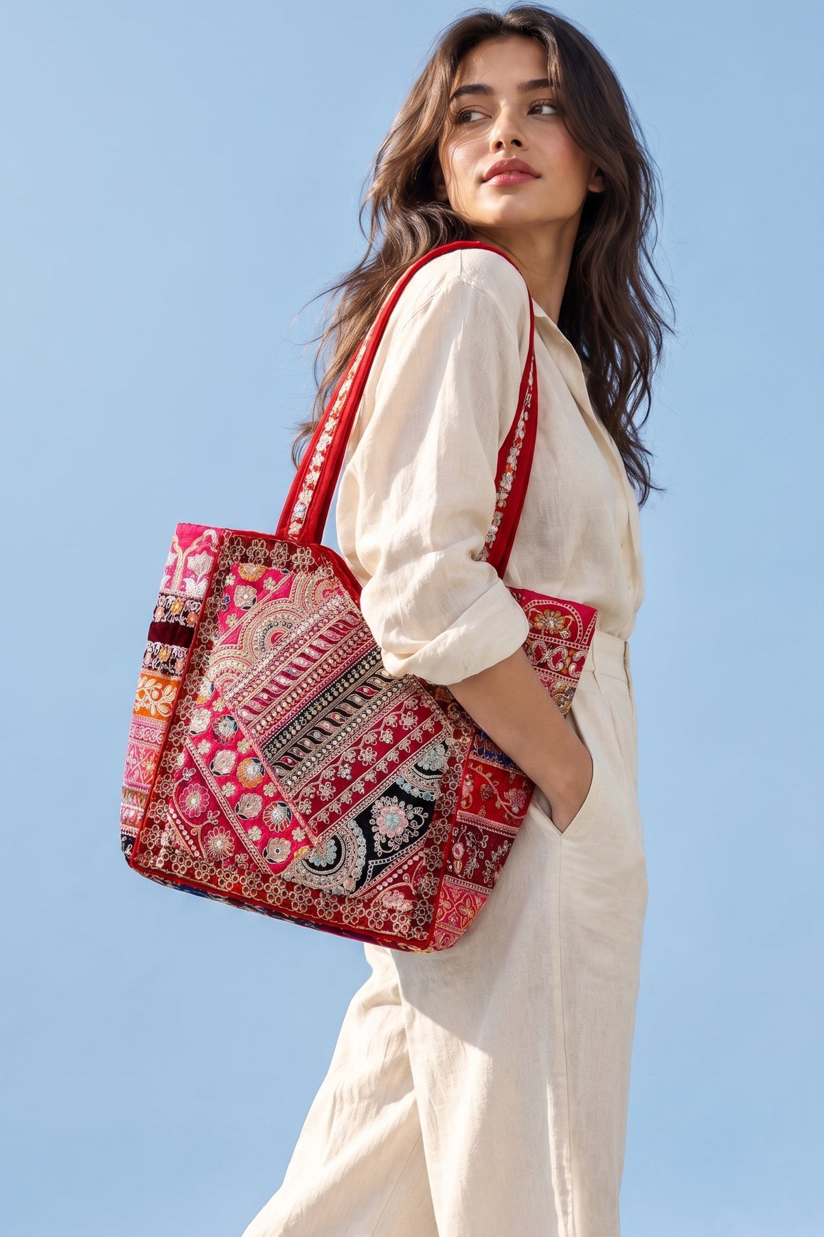A woman in a white dress carries a large red and multicolor patterned patchwork tote bag against a clear blue sky.