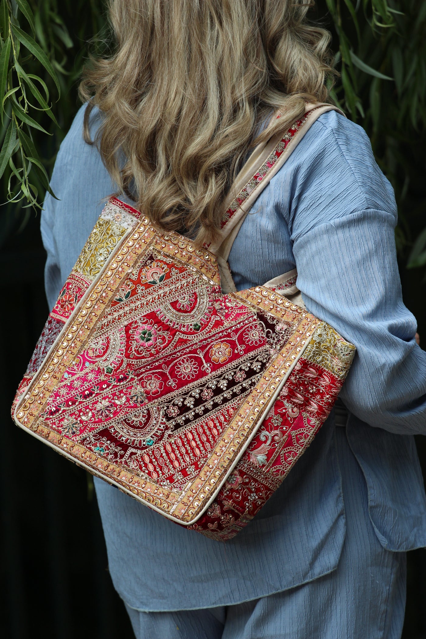 A woman wearing a light blue denim outfit carries a large, ornate patchwork tote bag with intricate red, gold, and beige embroidery over her shoulder.