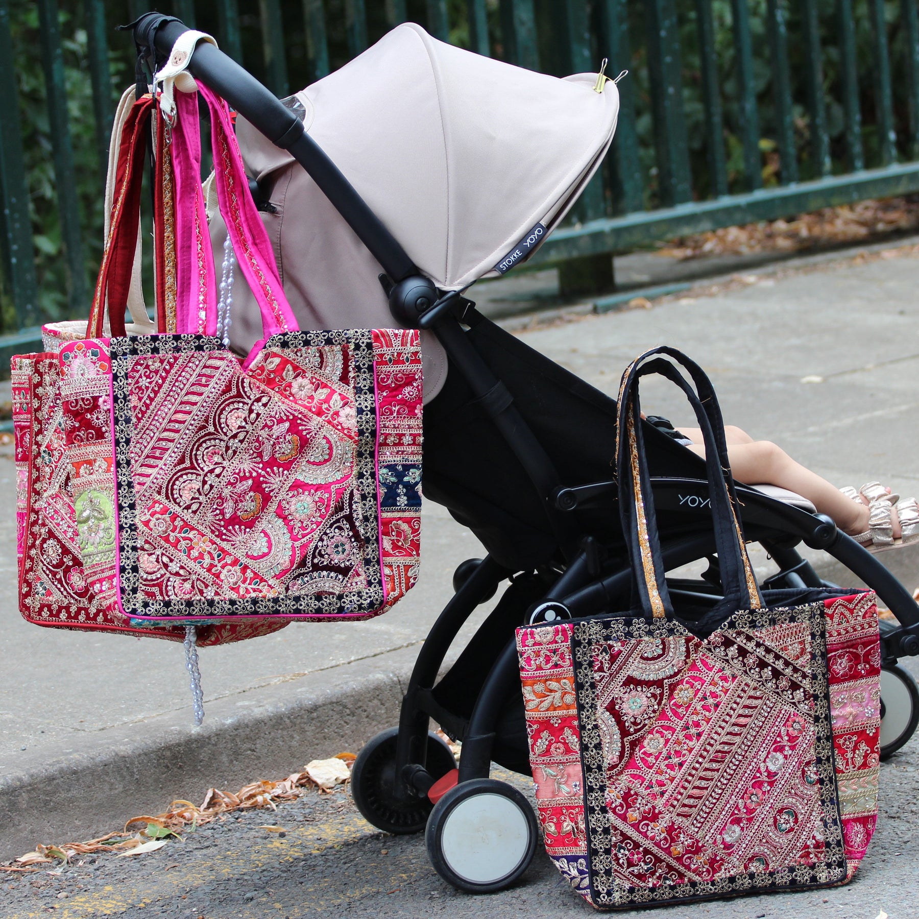 Two patterned tote bags with pink and red paisley and patchwork designs are shown in a real outdoor setting next to a black stroller on a sidewalk. One bag is hanging from the stroller handle, and the other is resting on the stroller seat.